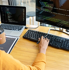 A person in an orange sweater codes on a desktop computer with a laptop nearby, holding a coffee cup at a wooden table.