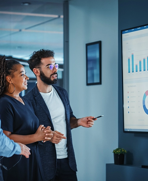 A group of professionals examines data on a large screen displaying graphs and charts in a modern office setting.