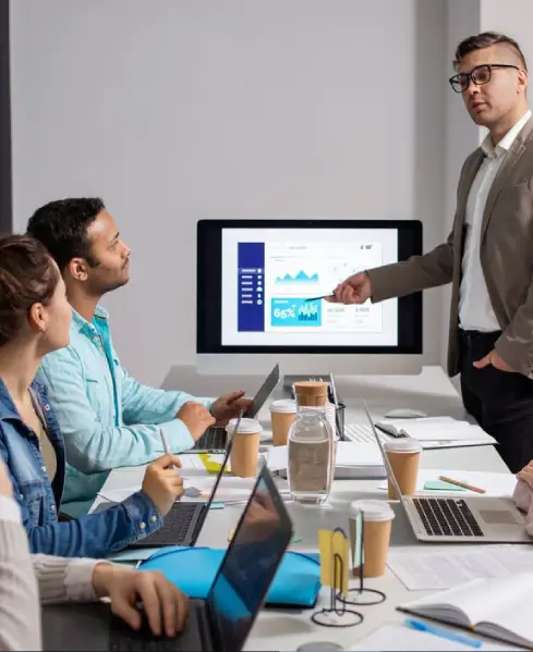 A diverse group of professionals collaborates in a modern meeting room, viewing a presentation on a computer screen. Laptops and coffee cups on the table.